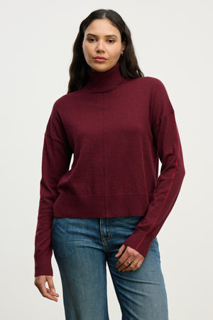 A woman wears the Velvet by Graham & Spencer LIBRA SWEATER in burgundy cotton cashmere with blue jeans, standing against a plain light background.