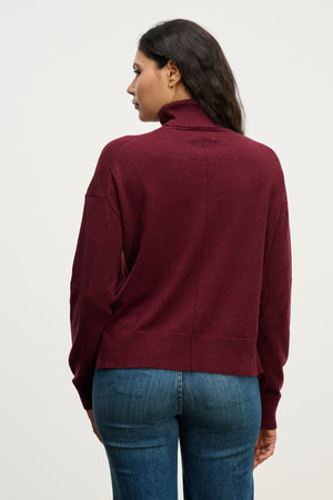 A woman faces away, wearing the LIBRA SWEATER by Velvet by Graham & Spencer in maroon with a boxy fit and turtleneck, paired with blue jeans against a plain light background.