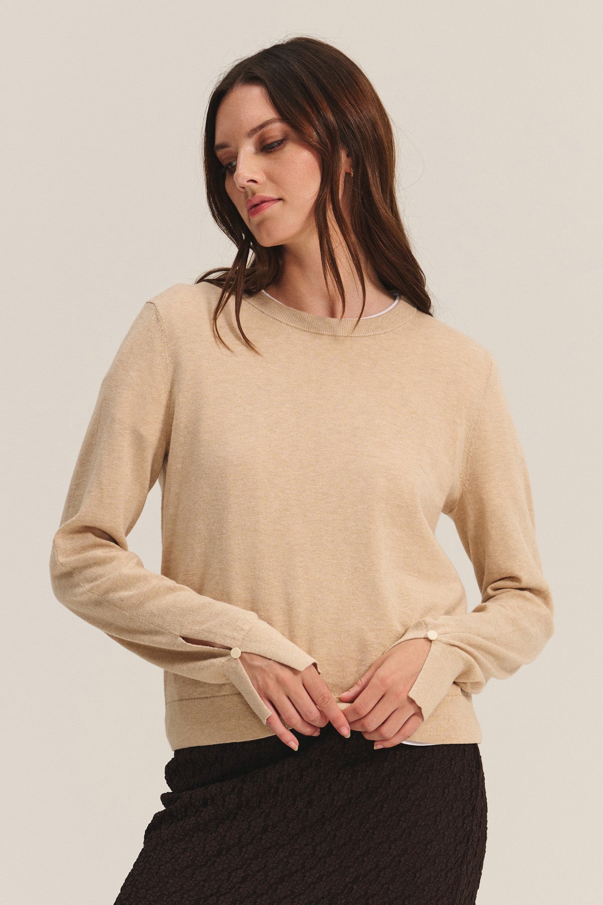 A woman wears the MELINA SWEATER by Velvet by Graham & Spencer, a beige cotton-cashmere crew neck with ribbed trims, paired with a textured dark skirt against a light background.-40681731522753