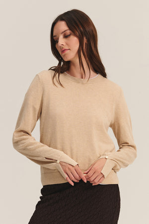 A woman wears the MELINA SWEATER by Velvet by Graham & Spencer, a beige cotton-cashmere crew neck with ribbed trims, paired with a textured dark skirt against a light background.