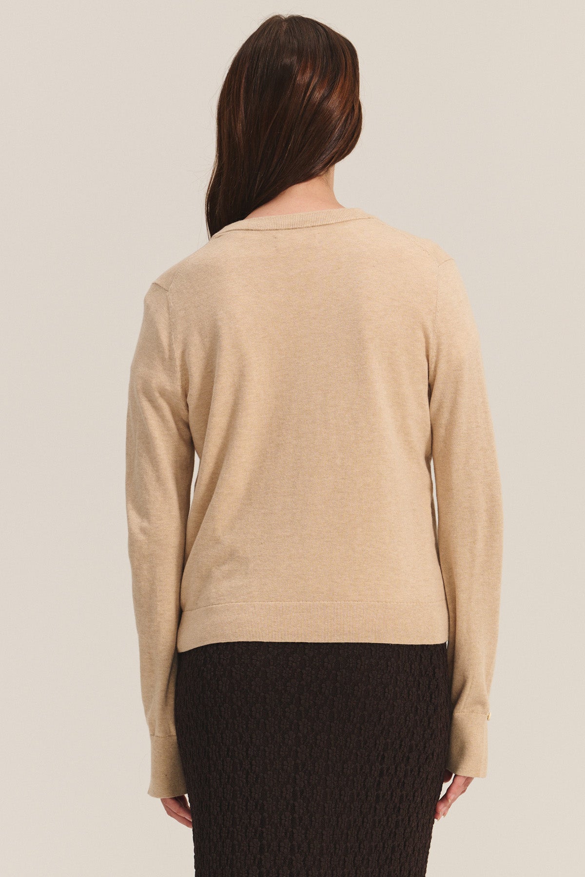 A person stands with their back to the camera, wearing Velvet by Graham & Spencer's MELINA SWEATER—a beige cotton-cashmere crew neck with ribbed trims—paired with a textured black skirt against a neutral background.-40681731489985