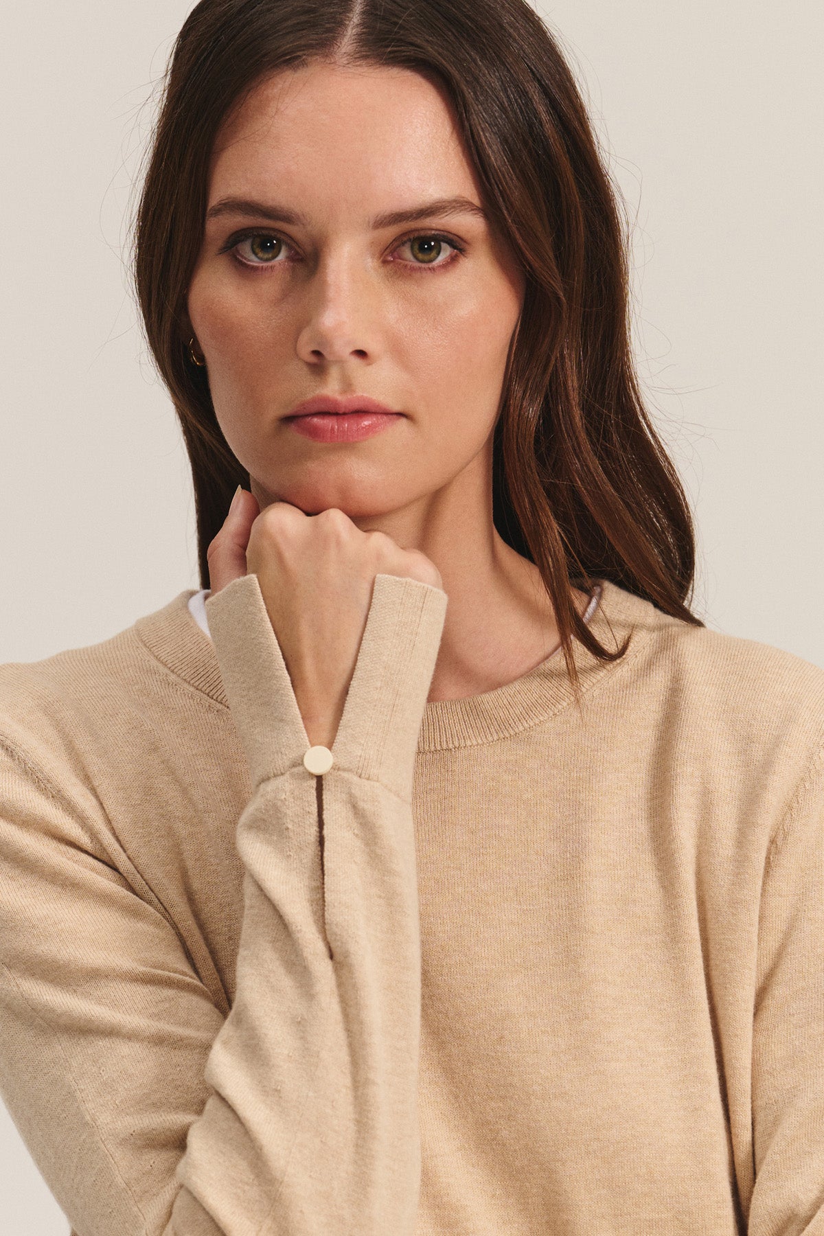 A woman with long brown hair wears the MELINA SWEATER by Velvet by Graham & Spencer—a beige cotton-cashmere crew neck with ribbed trims—and looks directly at the camera, chin resting on her fist.