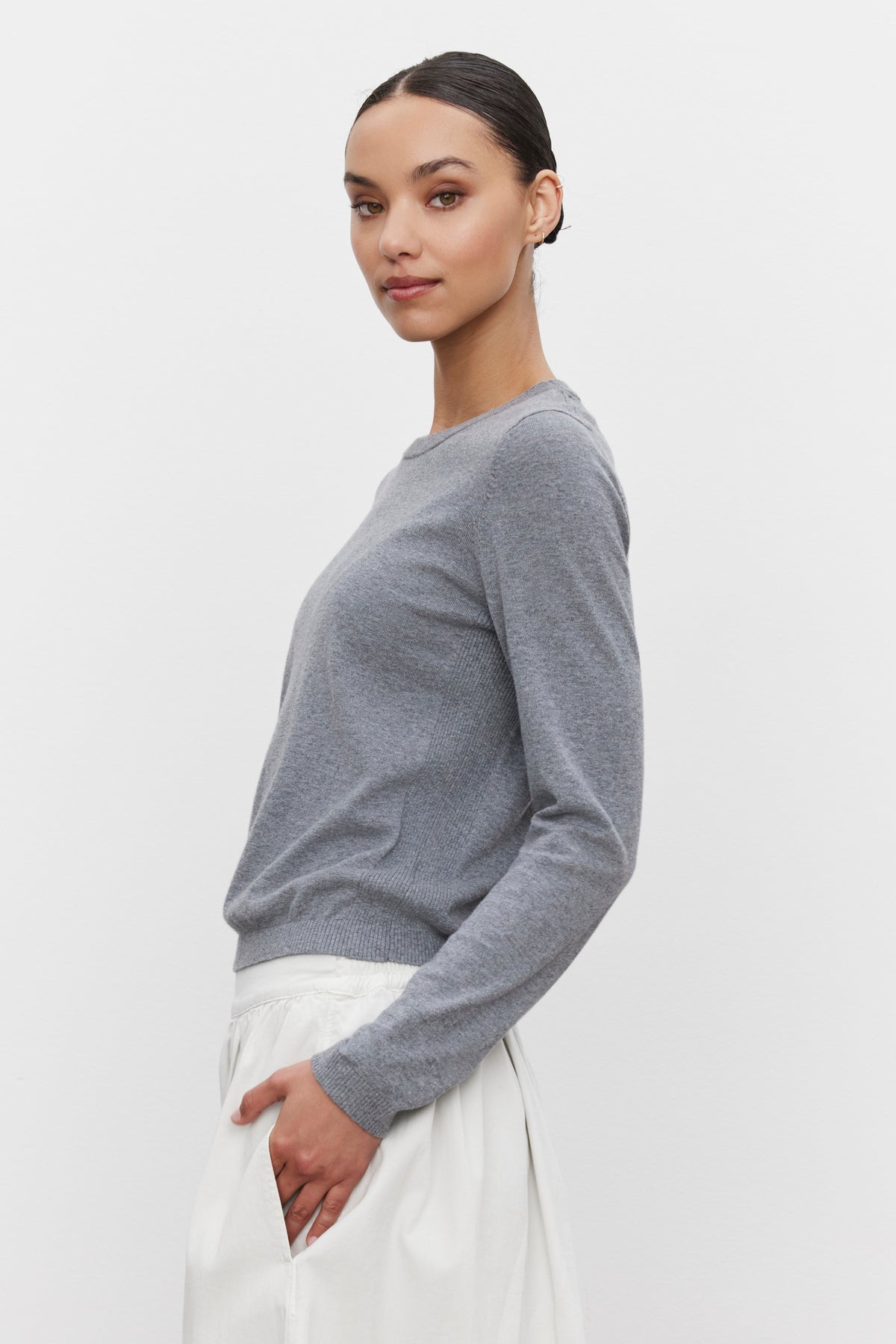   A person wearing the Velvet by Graham & Spencer ROXY SWEATER, a grey, long-sleeve, lightweight sweater with ribbed details, and white pants stands against a plain white background, looking at the camera with one hand in their pocket. 