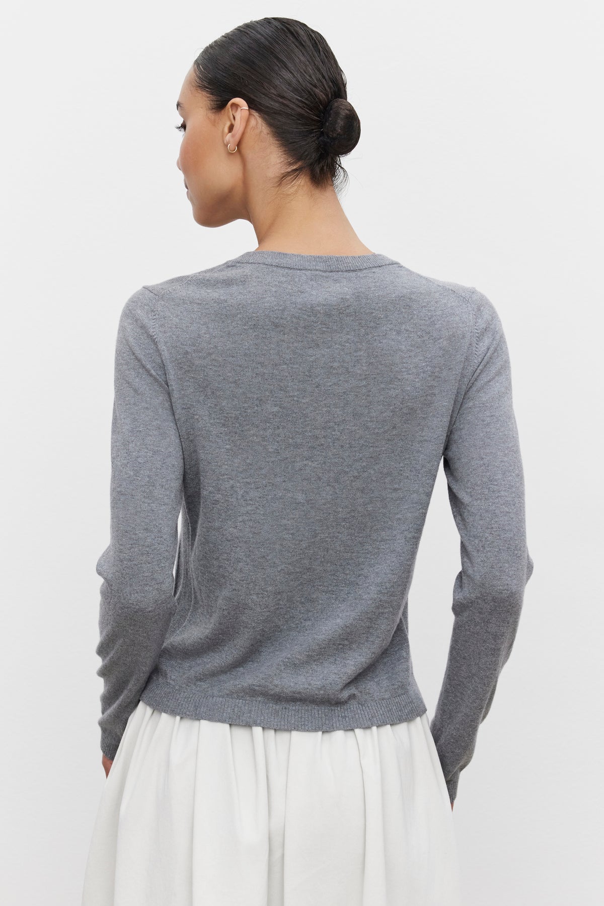   A person with dark hair in a bun, wearing the ROXY SWEATER in gray lux cotton cashmere with ribbed details by Velvet by Graham & Spencer, paired with a white skirt and photographed from the back. 