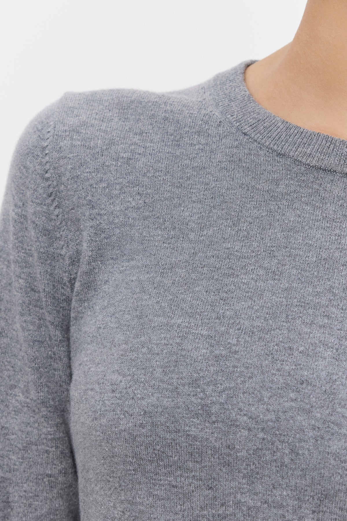   Close-up of a person wearing the ROXY SWEATER by Velvet by Graham & Spencer. The lightweight gray sweater features ribbed details on the long sleeves and round neck, and is made of soft lux cotton cashmere knit fabric. The background is plain and light-colored. 