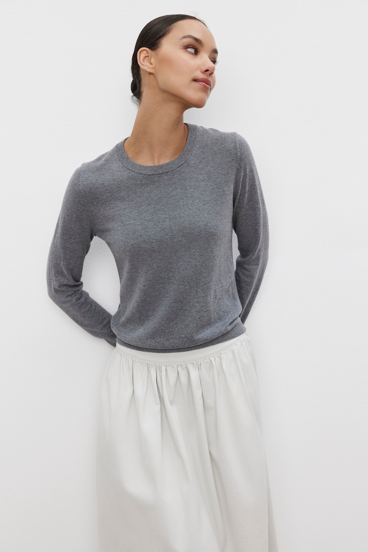   A person standing against a white background wears the Velvet by Graham & Spencer ROXY SWEATER in gray and a light-colored skirt, looking upwards with hands behind the back. 