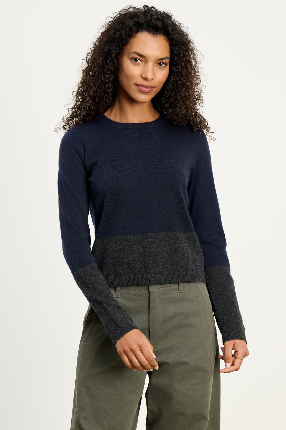 A woman wears the ROXY SWEATER by Velvet by Graham & Spencer—an ultra-soft cotton cashmere crewneck in navy and dark gray colorblock—paired with olive green pants, standing against a plain light background.-39742392205505