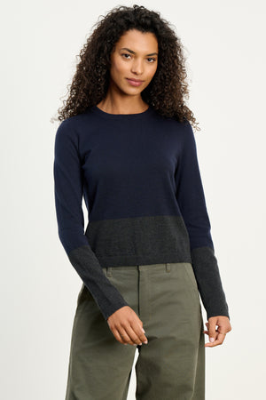 A woman wears the ROXY SWEATER by Velvet by Graham & Spencer—an ultra-soft cotton cashmere crewneck in navy and dark gray colorblock—paired with olive green pants, standing against a plain light background.