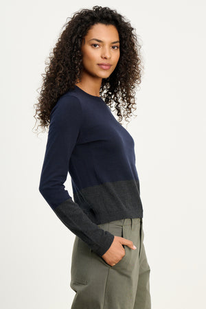 A woman with curly hair wears the ROXY SWEATER by Velvet by Graham & Spencer—a navy and dark gray lightweight crewneck—paired with olive green pants, standing against a plain light background.