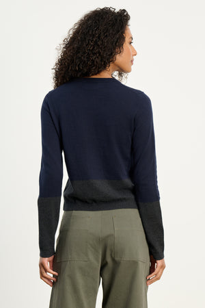 A person with curly hair is seen from the back, modeling the ROXY SWEATER by Velvet by Graham & Spencer—a navy and dark gray colorblock cotton-cashmere crewneck—paired with olive green pants against a plain light background.