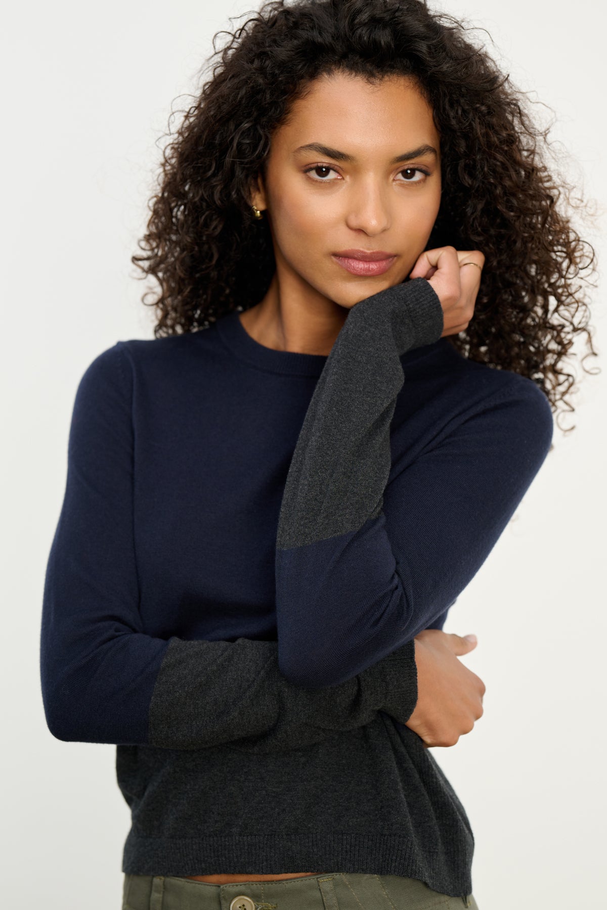 A woman with curly hair models the Velvet by Graham & Spencer ROXY SWEATER in navy blue and dark gray with a crew neckline, paired with olive green pants, standing against a plain light background.-39742392074433