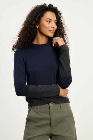 A woman with curly hair models the Velvet by Graham & Spencer ROXY SWEATER in navy and dark gray cotton-cashmere, featuring a crew neck, paired with olive green pants against a plain light background.