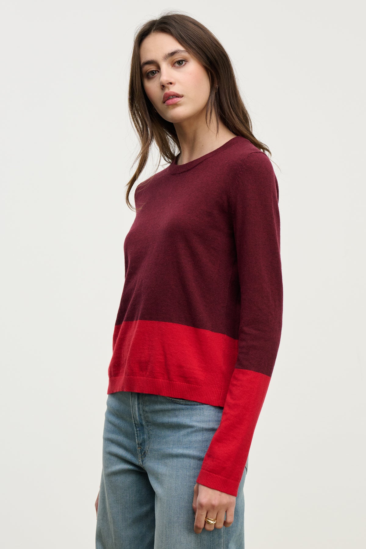 A woman wearing the ROXY SWEATER by Velvet by Graham & Spencer—a long-sleeve, burgundy and red color-block lightweight sweater—with blue jeans stands against a plain light background.-40046135312577