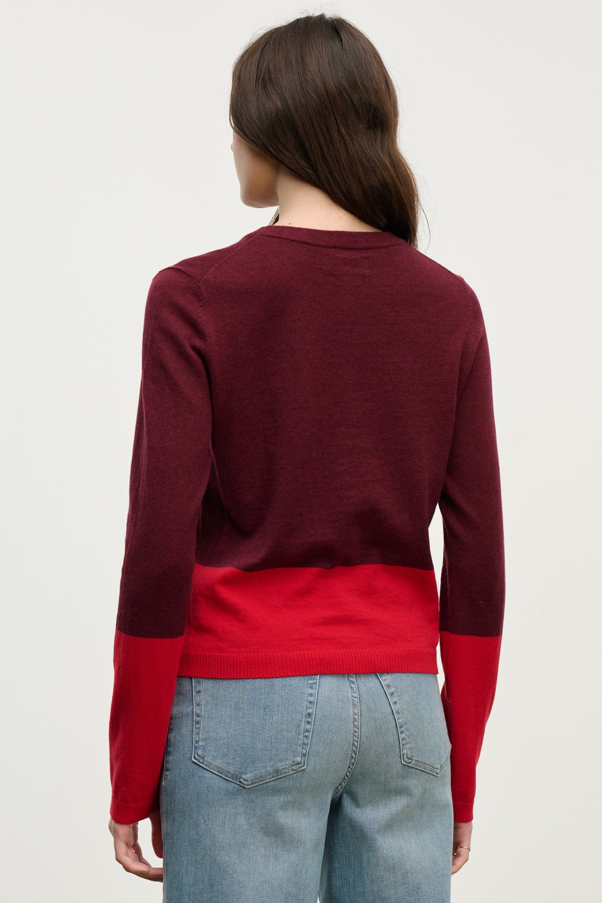 A person with long brown hair, seen from the back against a light background, wears blue jeans and a maroon-and-red ROXY SWEATER by Velvet by Graham & Spencer. The lightweight sweater adds comfort and style to the casual outfit.-40046135279809