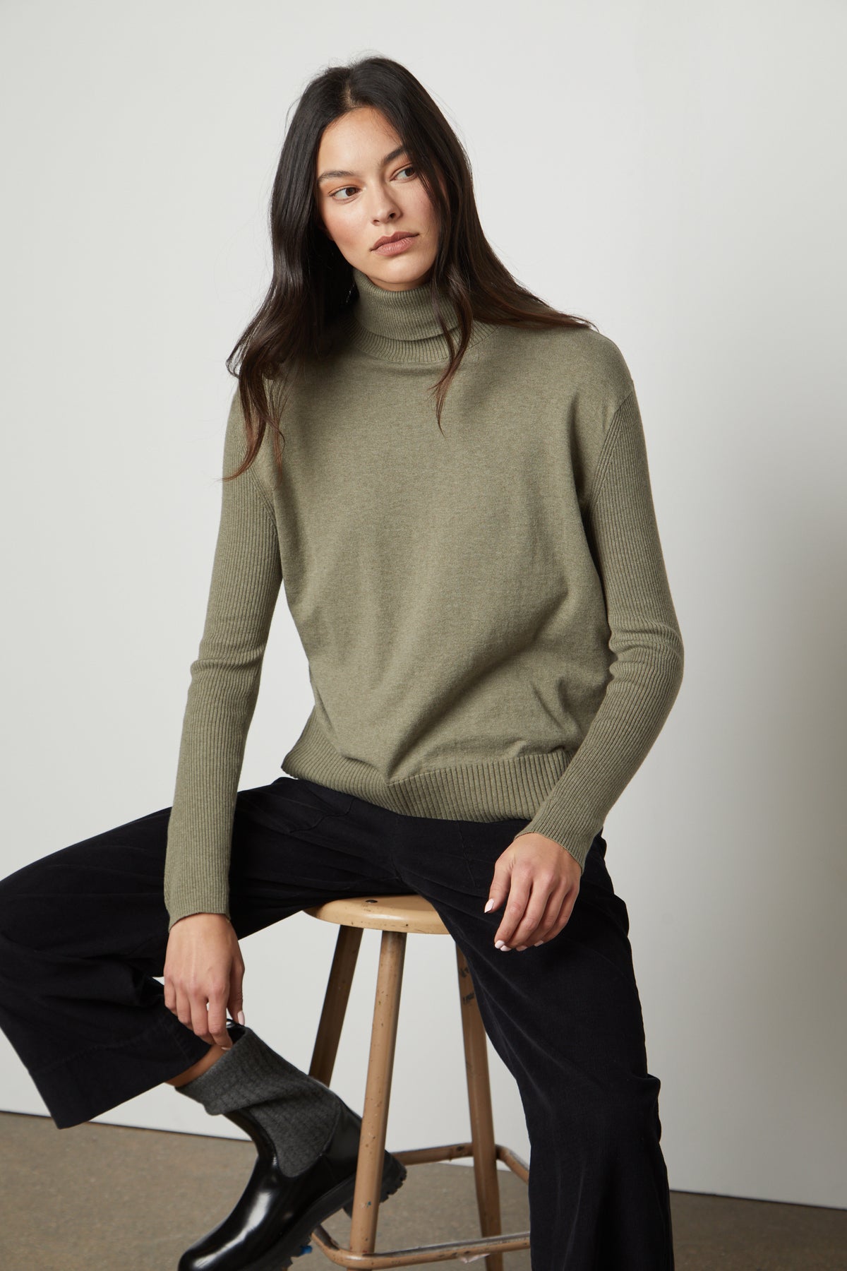   A woman is sitting on a stool wearing a fitted silhouette green SALLY MOCK NECK SWEATER by Velvet by Graham & Spencer. 