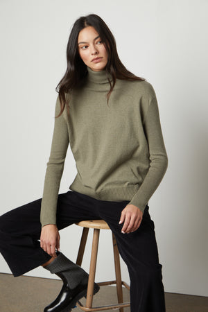A woman is sitting on a stool wearing a fitted silhouette green SALLY MOCK NECK SWEATER by Velvet by Graham & Spencer.