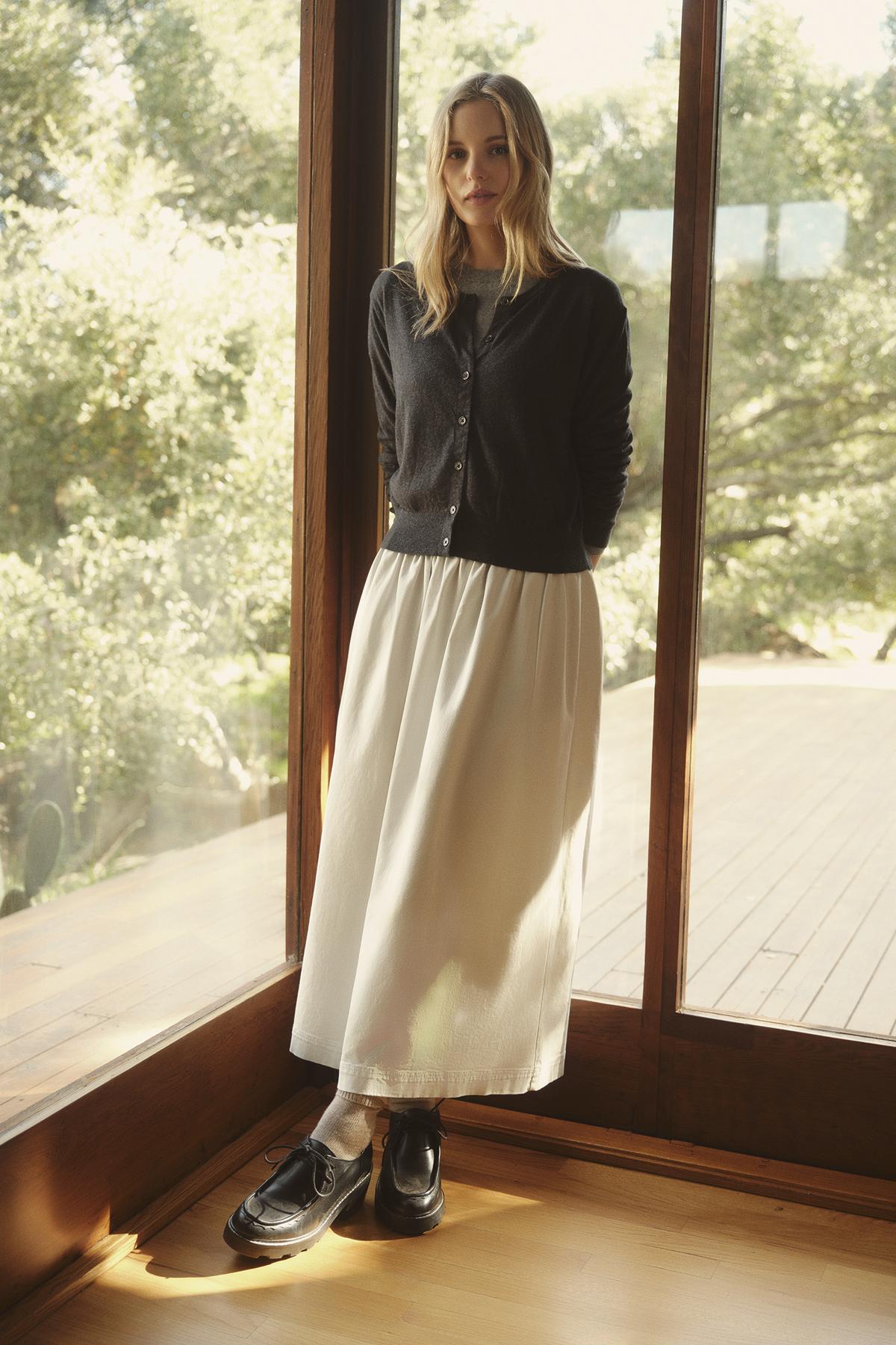   A person stands by a large window wearing a VIOLETTE CARDIGAN from Velvet by Graham & Spencer, paired with a long white skirt and black shoes. Sunlight streams through the window, illuminating the indoor room and the greenery outside. 