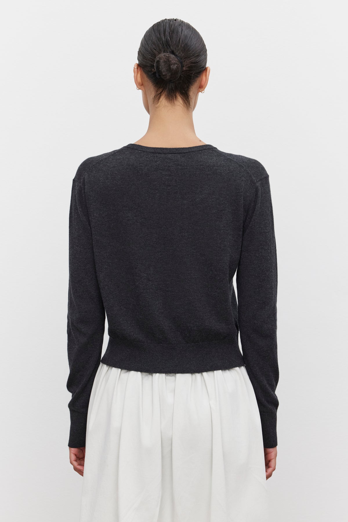 A person with dark hair in a bun stands facing away, wearing a long-sleeve black top and a white skirt, topped with the lightweight VIOLETTE CARDIGAN from Velvet by Graham & Spencer.-37676236243137