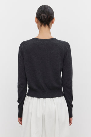 A person with dark hair in a bun stands facing away, wearing a long-sleeve black top and a white skirt, topped with the lightweight VIOLETTE CARDIGAN from Velvet by Graham & Spencer.