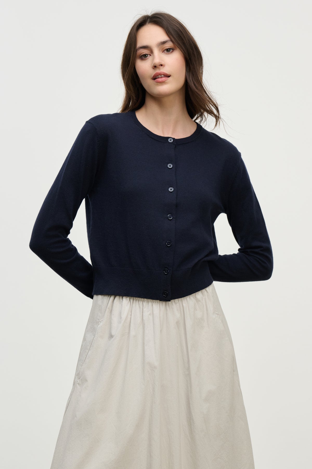 A woman wearing the VIOLETTE CARDIGAN by Velvet by Graham & Spencer in navy blue and a light beige skirt stands against a plain, light background.-39800434688193