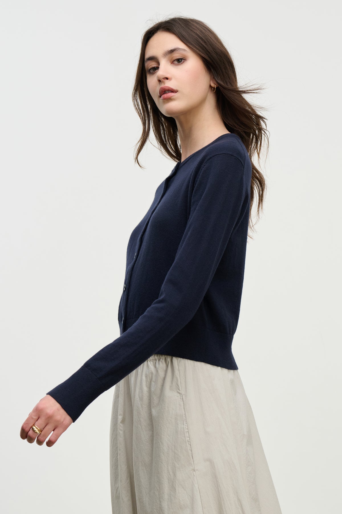 Young woman with long brown hair wears the VIOLETTE CARDIGAN by Velvet by Graham & Spencer, navy and cropped, paired with a light beige skirt. She stands sideways against a plain light background.-39800434655425