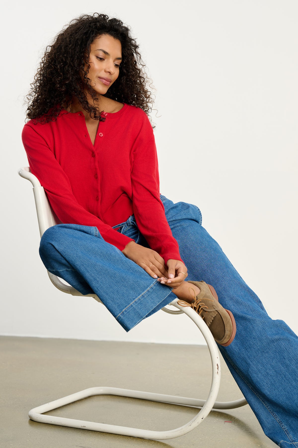 A woman with curly hair sits on a white chair, wearing the VIOLETTE CARDIGAN by Velvet by Graham & Spencer, blue wide-leg jeans, and brown shoes against a plain background.-39800434327745