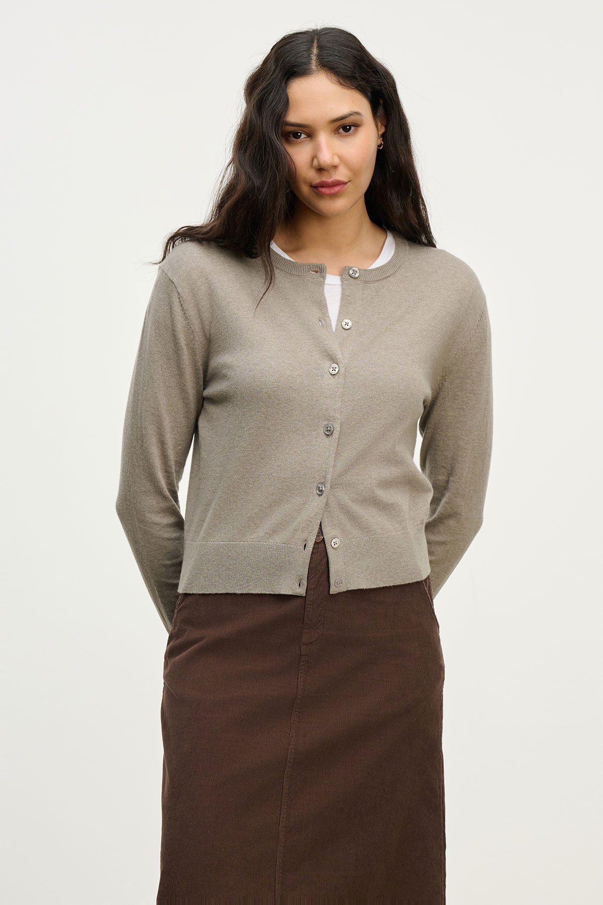   A woman faces the camera wearing the VIOLETTE CARDIGAN by Velvet by Graham & Spencer over a white top and brown skirt, with her hands behind her back against a plain light background. 