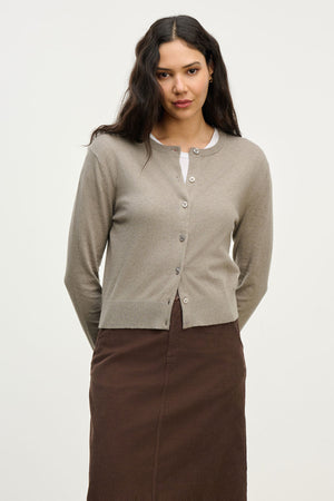 A woman faces the camera wearing the VIOLETTE CARDIGAN by Velvet by Graham & Spencer over a white top and brown skirt, with her hands behind her back against a plain light background.