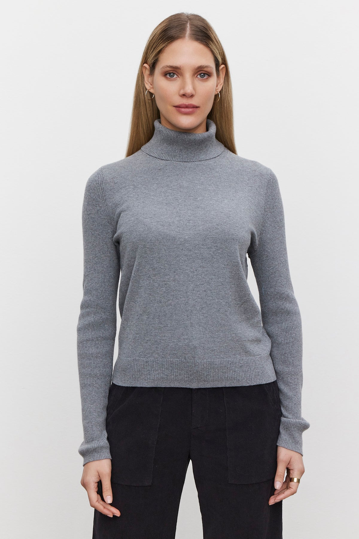   A person with long, straight hair is wearing the KORIE TURTLENECK SWEATER by Velvet by Graham & Spencer, featuring ribbed details, paired with dark pants, standing against a plain white background. 