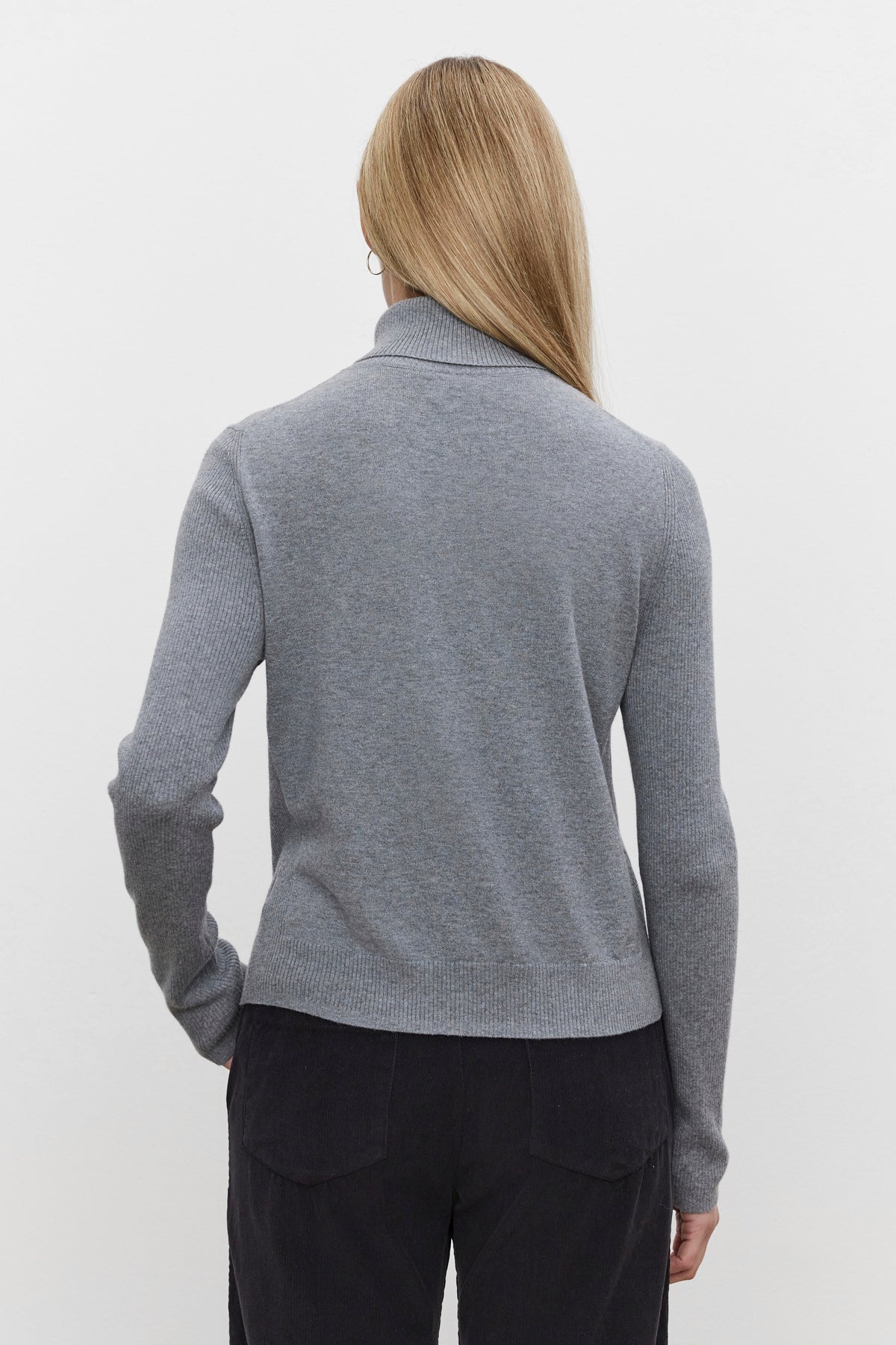   A person with long hair, seen from behind, is wearing a luxurious KORIE TURTLENECK SWEATER by Velvet by Graham & Spencer. This gray long-sleeve turtleneck sweater is made of cotton cashmere and dark pants, featuring ribbed details for added texture. 