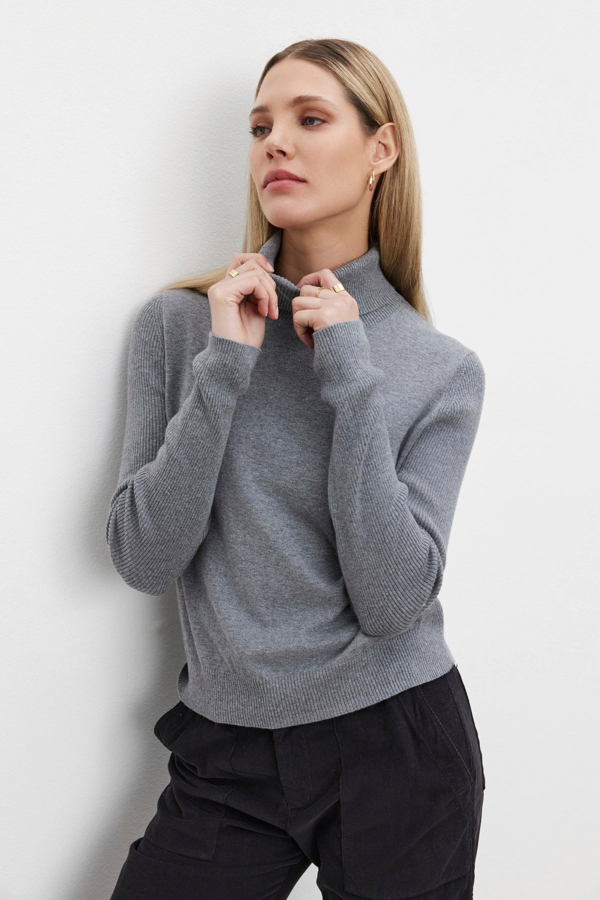   A woman with long hair wears the KORIE TURTLENECK SWEATER in light gray from Velvet by Graham & Spencer, paired with dark pants, as she stands against a plain white background. 