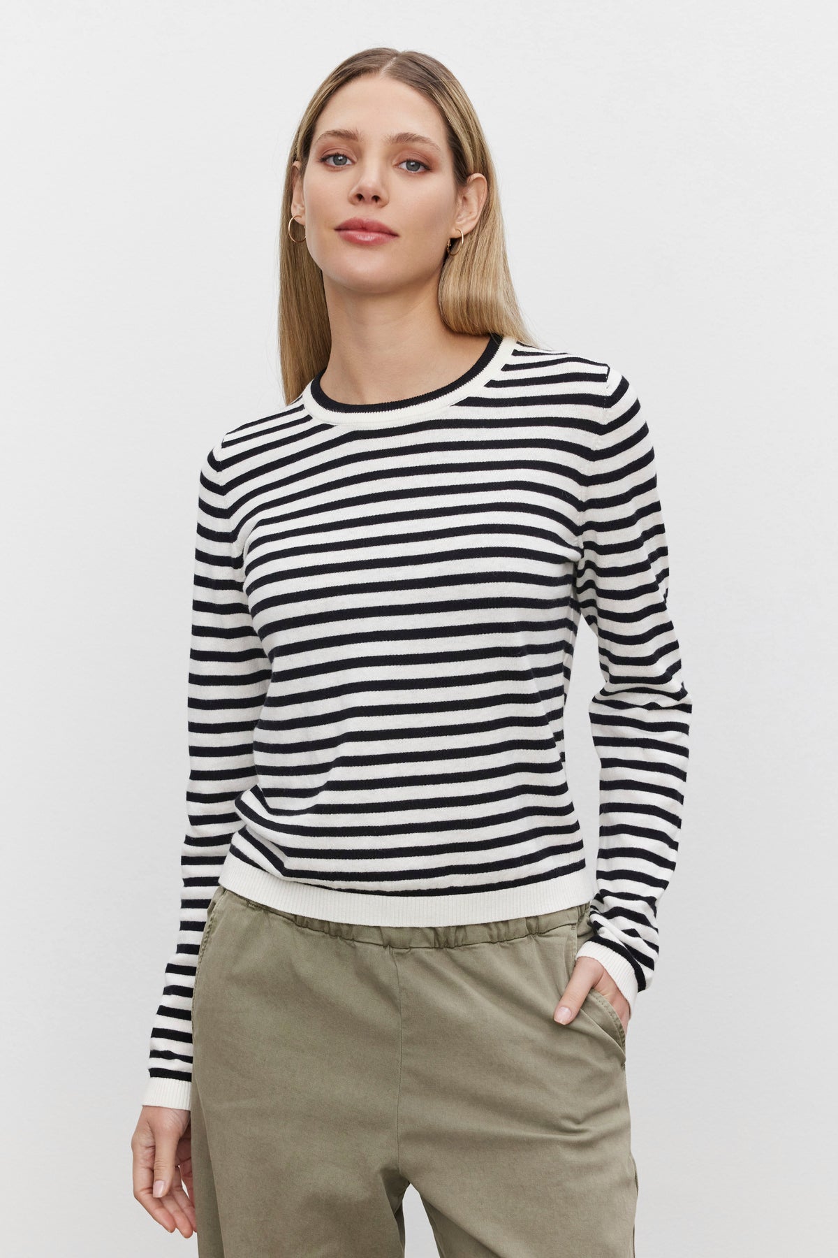   A person stands wearing a black and white striped ROXY SWEATER by Velvet by Graham & Spencer and khaki pants, with one hand in their pocket against a plain white background. 