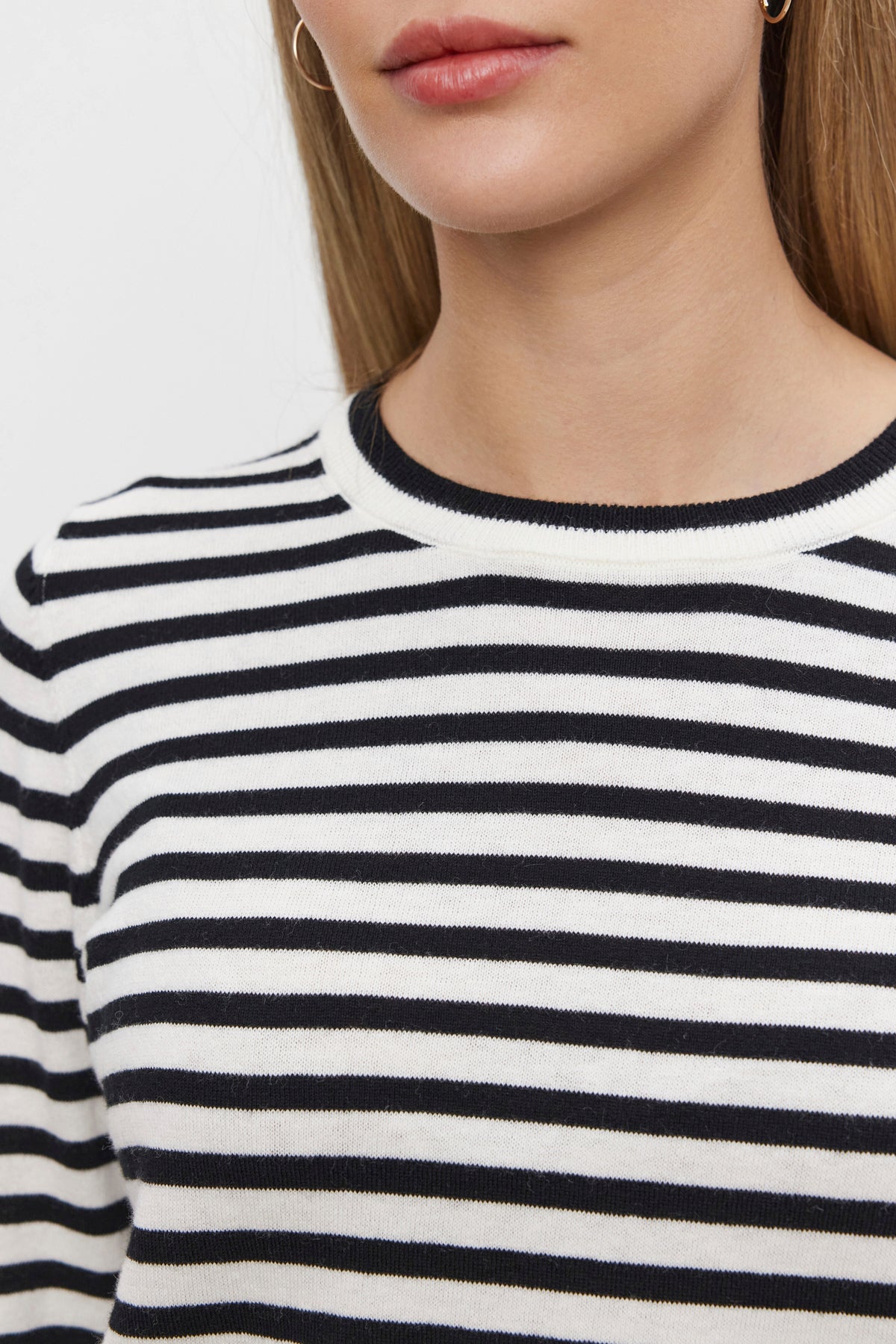   A close-up image shows a person wearing the ROXY SWEATER by Velvet by Graham & Spencer, featuring ribbed details and a black-and-white striped pattern. The round neckline frames the lower half of the face, neck, and upper torso, hinting at the softness of its luxurious cotton-cashmere fabric. 