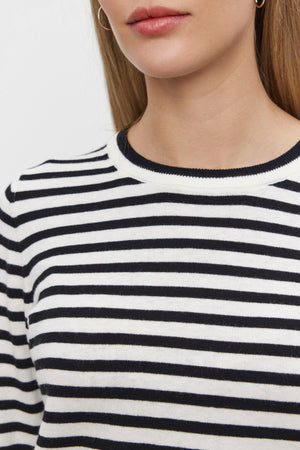 A close-up image shows a person wearing the ROXY SWEATER by Velvet by Graham & Spencer, featuring ribbed details and a black-and-white striped pattern. The round neckline frames the lower half of the face, neck, and upper torso, hinting at the softness of its luxurious cotton-cashmere fabric.