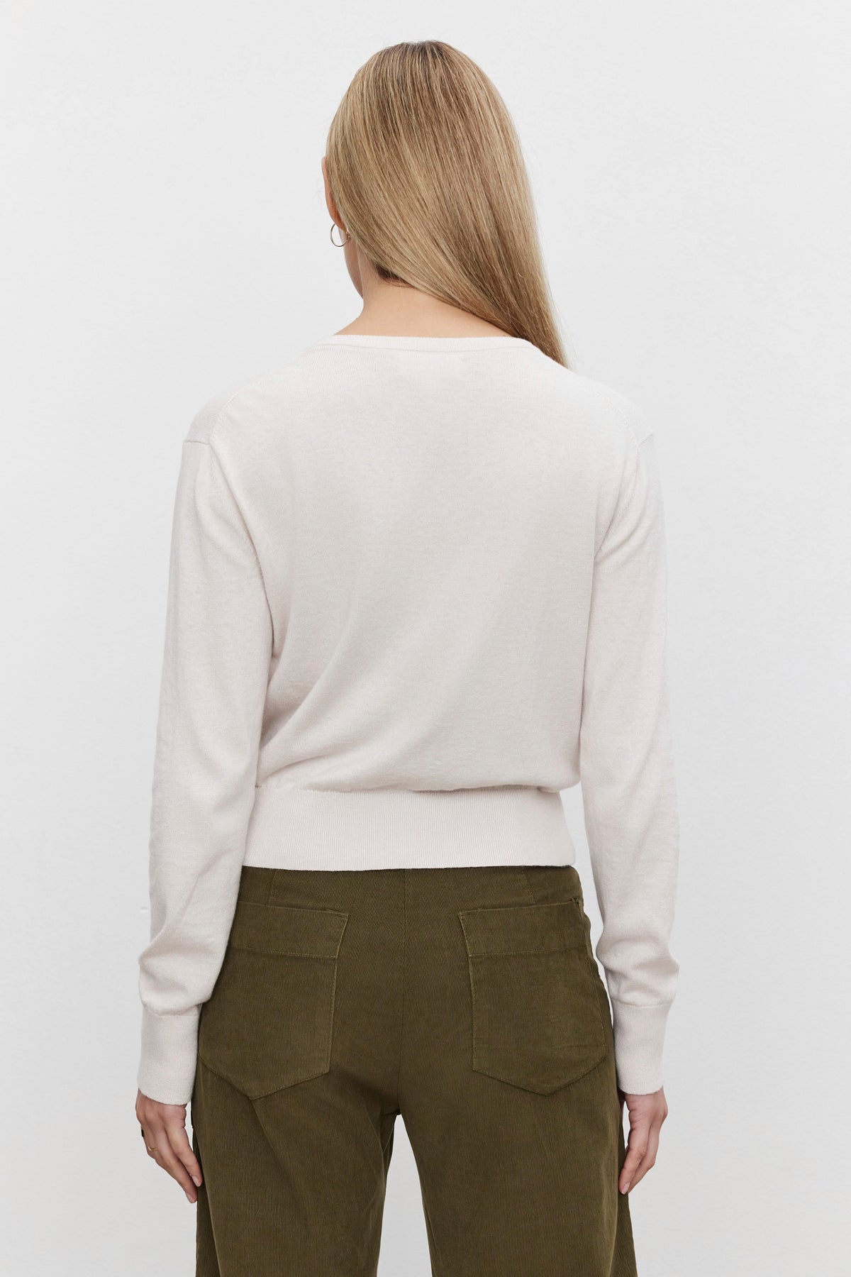 A person with long, blonde hair is shown from the back, wearing the VIOLETTE CARDIGAN by Velvet by Graham & Spencer in a light-colored, cropped fit and olive green pants, exuding a lightweight feel.-37676917817537