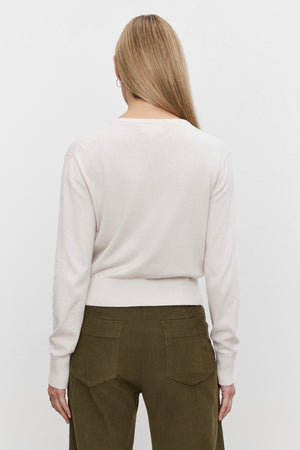 A person with long, blonde hair is shown from the back, wearing the VIOLETTE CARDIGAN by Velvet by Graham & Spencer in a light-colored, cropped fit and olive green pants, exuding a lightweight feel.