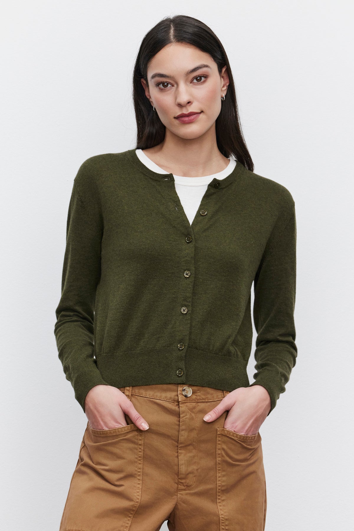   A woman with long dark hair, wearing the Velvet by Graham & Spencer Violette Cardigan in green, which features a lux cotton cashmere blend and a cropped fit over a white shirt, poses with her hands in the pockets of her tan pants against a plain white background. 