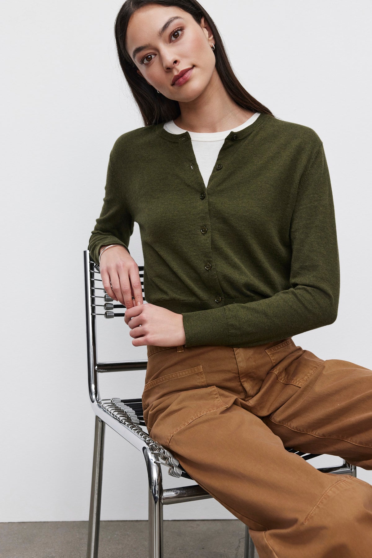   A person with long dark hair sits on a metal chair wearing the cropped fit VIOLETTE CARDIGAN by Velvet by Graham & Spencer in olive green, along with a white shirt and brown pants against a plain background. 