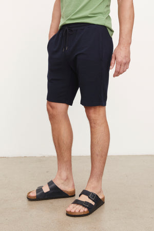 A man wearing a green shirt and navy ATLAS LUXE FLEECE DRAWSTRING SHORTS with a cozy interior, by Velvet by Graham & Spencer.