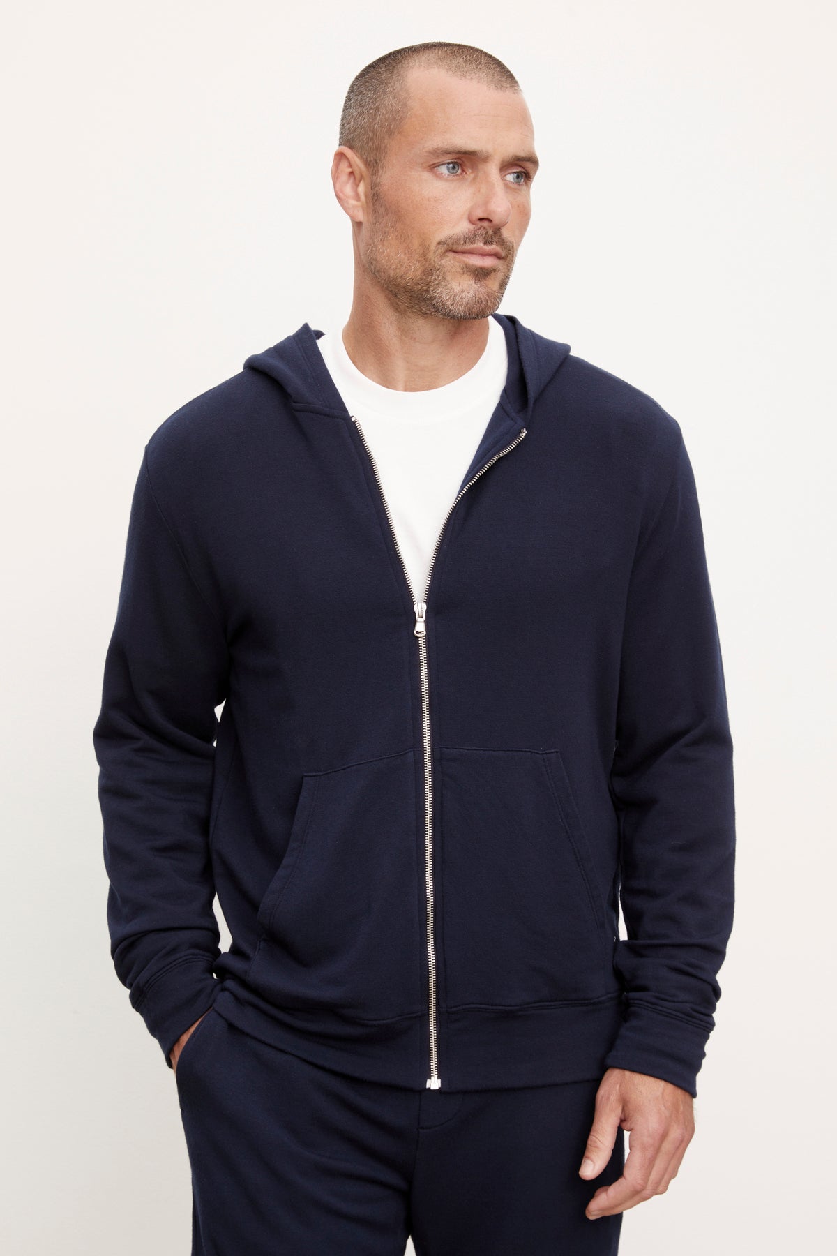   A man wearing a RODAN LUXE FLEECE ZIP HOODIE navy blue hoodie from Velvet by Graham & Spencer over a white t-shirt, standing against a neutral background, looking to his left. 