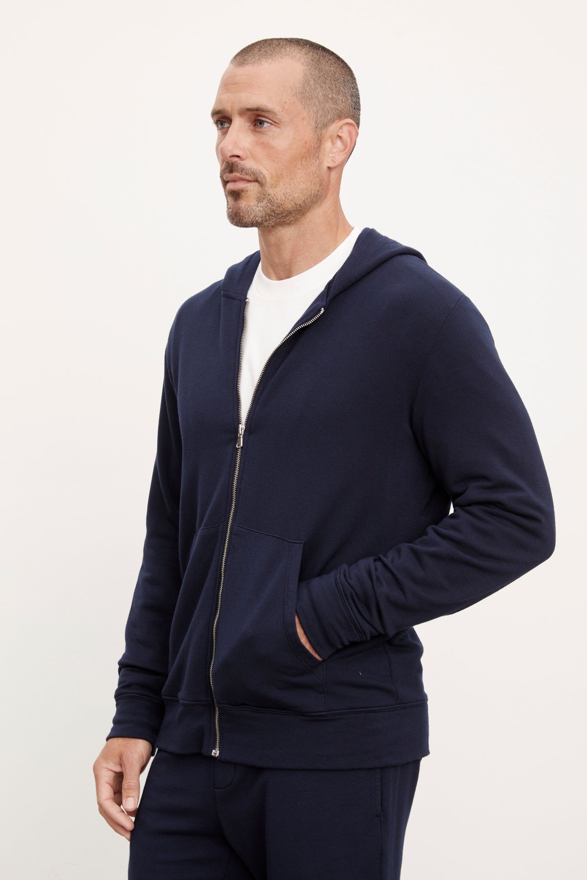 A man in a navy blue non-bulky fit RODAN LUXE FLEECE ZIP HOODIE and matching pants, standing with his hand on his hip, looking to his left by Velvet by Graham & Spencer.-36731041349825