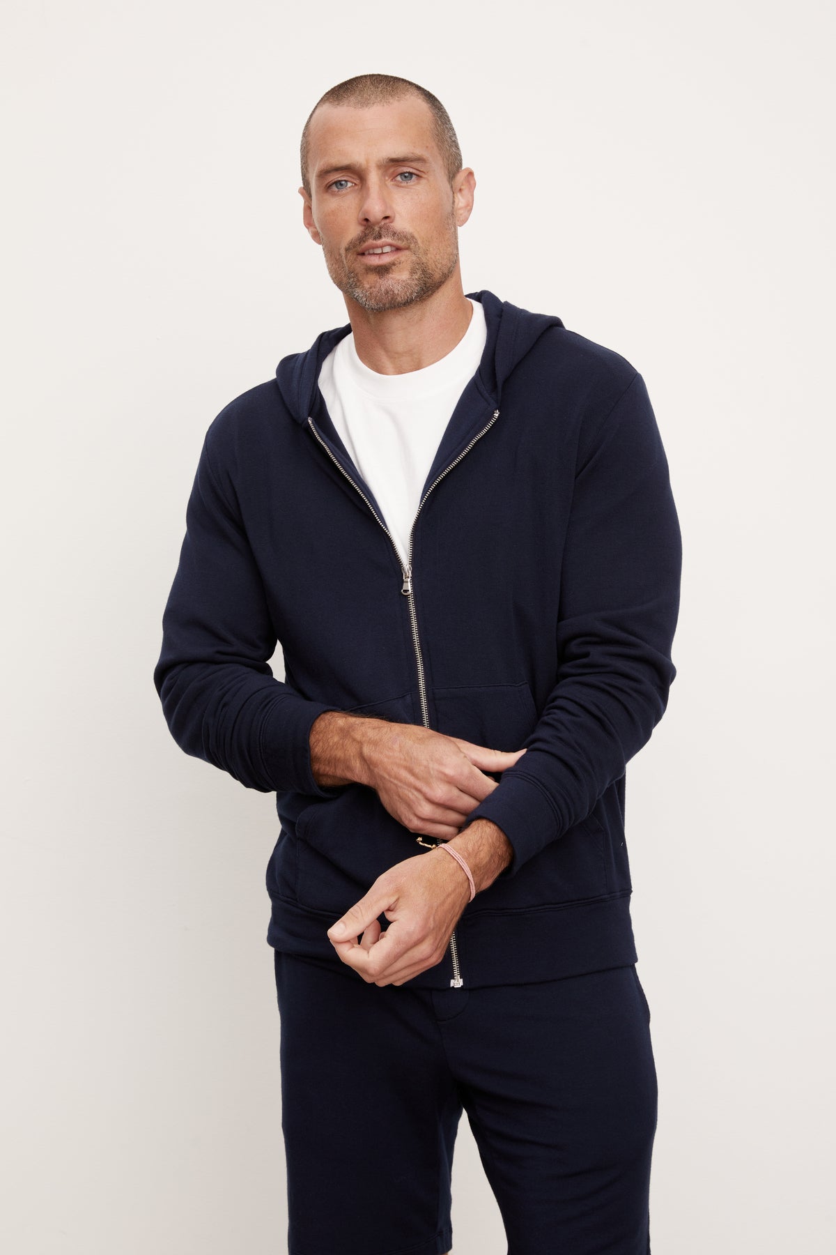 Man standing against a white background wearing a navy blue, medium-weight RODAN LUXE FLEECE ZIP HOODIE hoodie and matching pants, with hands partially in pockets.-36731041415361
