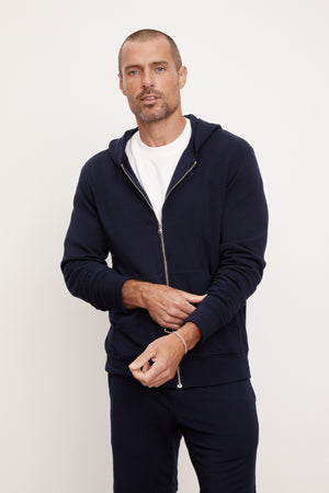 Man standing against a white background wearing a navy blue, medium-weight RODAN LUXE FLEECE ZIP HOODIE hoodie and matching pants, with hands partially in pockets.