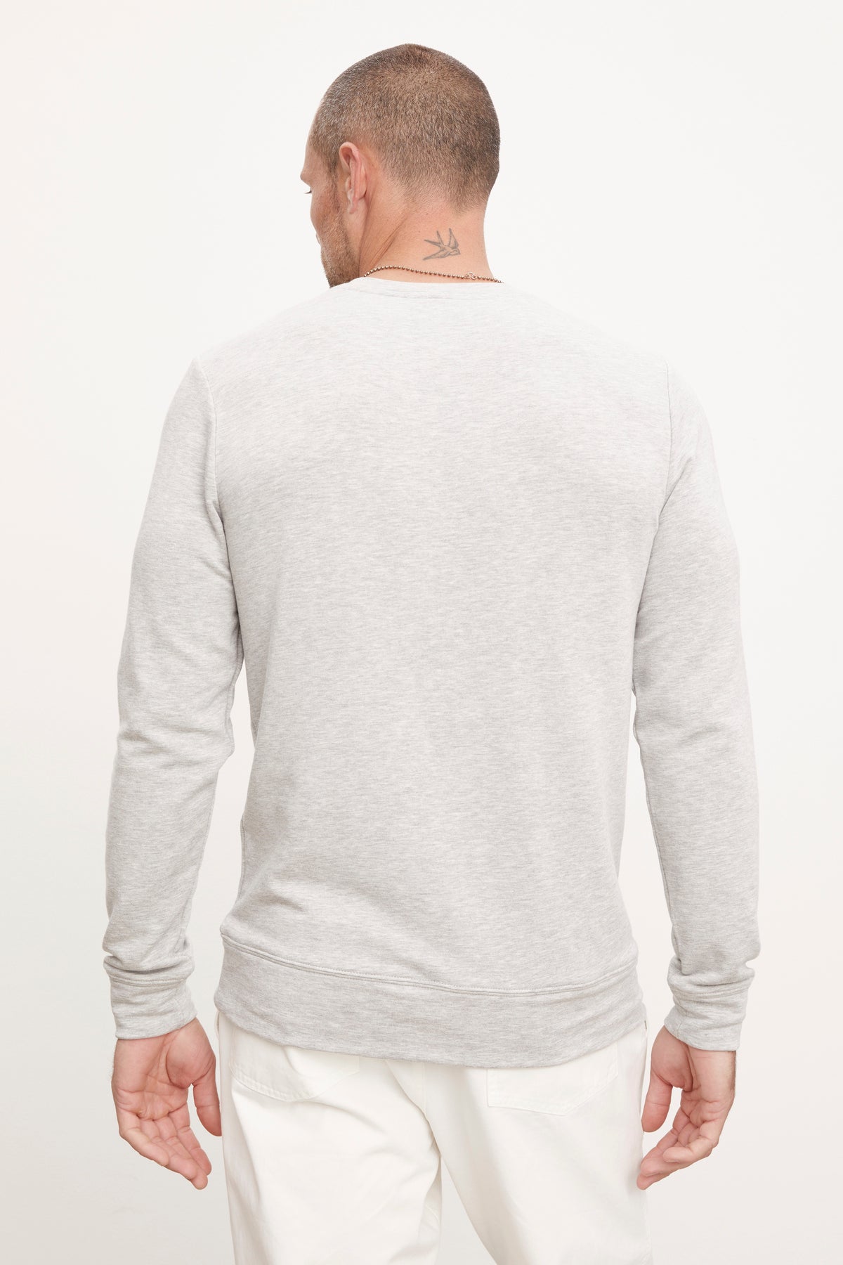   A person with short hair facing away is dressed in the SOREN PULLOVER, a gray long-sleeve shirt from Velvet by Graham & Spencer, made of medium-weight fleece, paired with white pants and featuring a small neck tattoo. 