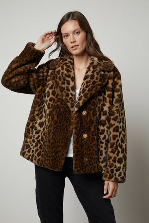 A woman wears a Velvet by Graham & Spencer AMANI LEOPARD LUX FAUX FUR JACKET, perfect for a fall wardrobe, and poses against a plain background. She has one hand raised to her head.