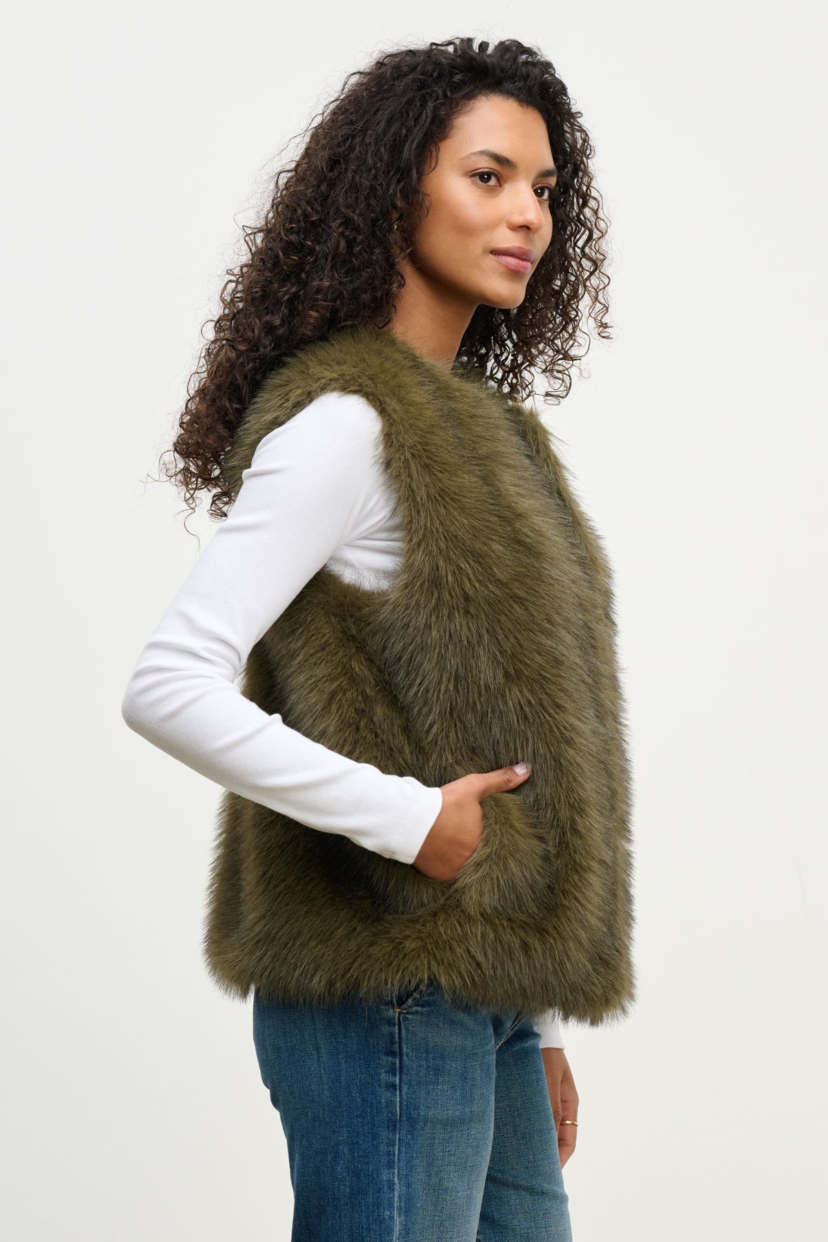   A woman with curly hair models the ANASTASIA FAUX FUR VEST by Velvet by Graham & Spencer, layered over a white long-sleeve shirt and blue jeans—a chic, cozy addition to any cold-weather wardrobe. 