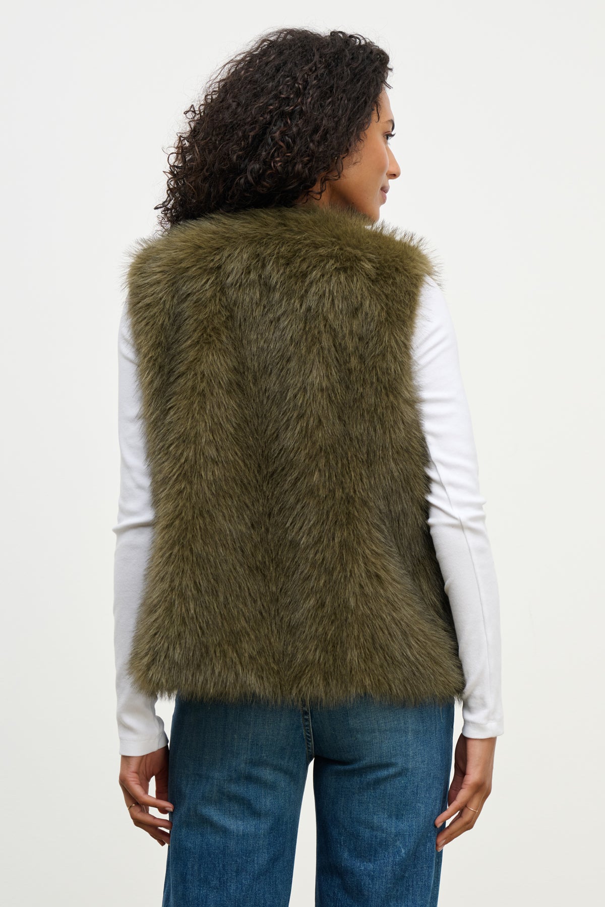   A woman faces away, wearing the ANASTASIA FAUX FUR VEST by Velvet by Graham & Spencer over a white long-sleeve shirt and blue jeans—a chic cold-weather essential shown against a plain background. 