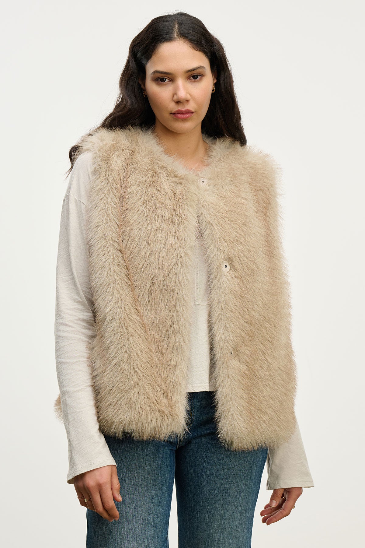 A woman wears the ANASTASIA FAUX FUR VEST by Velvet by Graham & Spencer over a long-sleeve shirt and blue jeans, standing against a light background—an ideal cozy luxury piece for your cold-weather wardrobe.-39815139688641