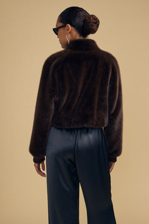 A person seen from behind, wearing a dark brown ARIANNA FAUX FUR BOMBER JACKET in a cropped, plush style with black trousers.