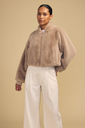 A person wearing a tan, zip-front ARIANNA FAUX FUR BOMBER JACKET by Velvet by Graham & Spencer and white wide-leg trousers.